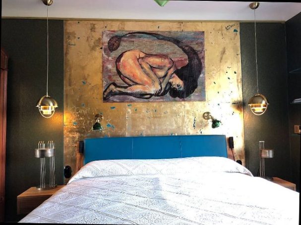 Schlafzimmer mit blauem Kopfteil, goldener Wandkunst und modernen Lampen.